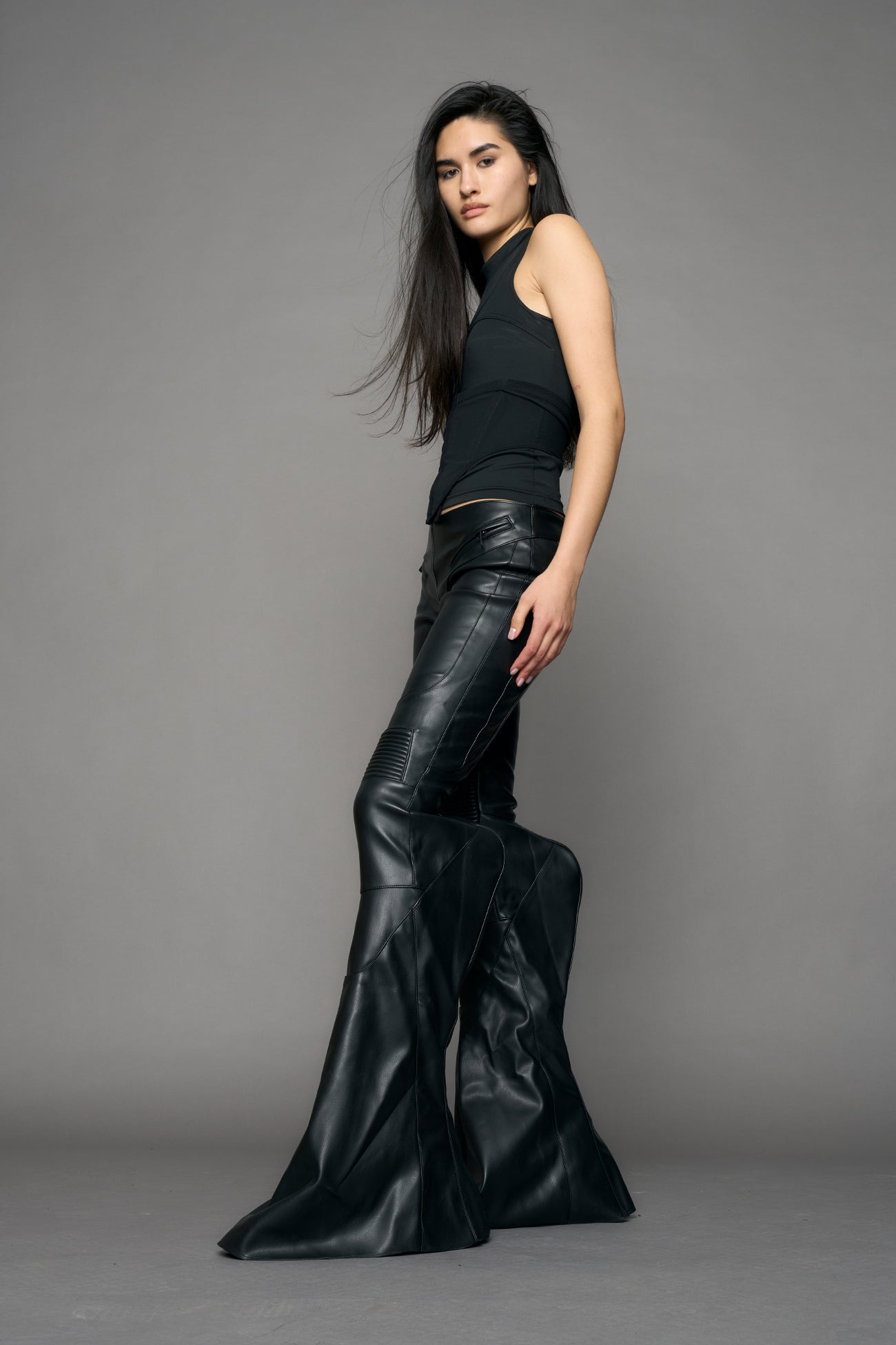 Black vegan leather stretch mutant pants with high waist, ribbed knee panels, and exaggerated flared legs by Namilia