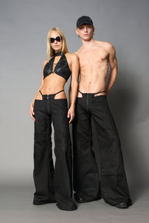 Low-rise flared pants in black cotton twill with bold side cut-outs and thong strap detailing. Featuring moto-style paneling, front zip closure, and a sculpted fit. A statement piece for rave, nightlife, and runway-inspired looks.