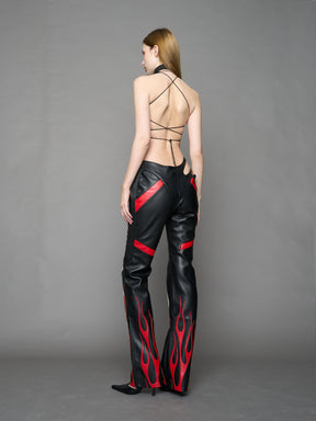 Model wears black/red vegan leather flare trousers with motocross paneling, padded details, flame and cross stitching, slogan embroidery, and an asymmetric waistband with adjustable elastic thong straps.