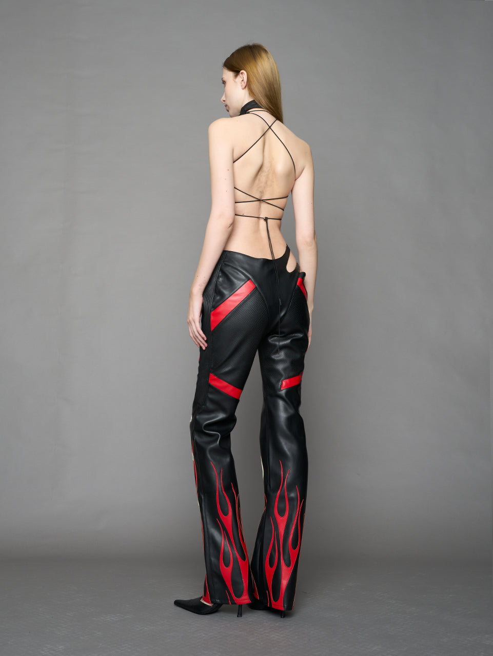Model wears black/red vegan leather flare trousers with motocross paneling, padded details, flame and cross stitching, slogan embroidery, and an asymmetric waistband with adjustable elastic thong straps.