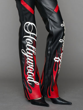 Model wears black/red vegan leather flare trousers with motocross paneling, padded details, flame and cross stitching, slogan embroidery, and an asymmetric waistband with adjustable elastic thong straps.