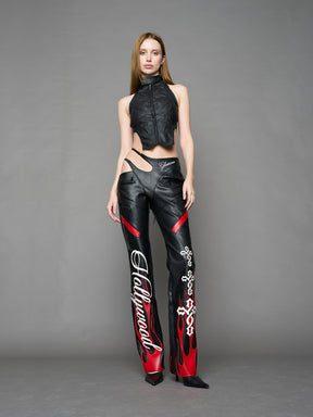 Model wears black/red vegan leather flare trousers with motocross paneling, padded details, flame and cross stitching, slogan embroidery, and an asymmetric waistband with adjustable elastic thong straps.