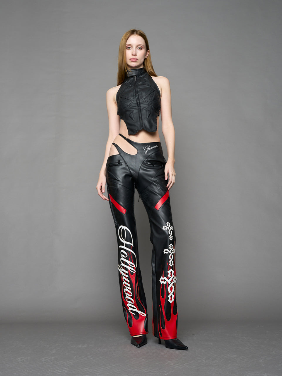 Model wears black/red vegan leather flare trousers with motocross paneling, padded details, flame and cross stitching, slogan embroidery, and an asymmetric waistband with adjustable elastic thong straps.