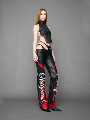 Model wears black/red vegan leather flare trousers with motocross paneling, padded details, flame and cross stitching, slogan embroidery, and an asymmetric waistband with adjustable elastic thong straps.