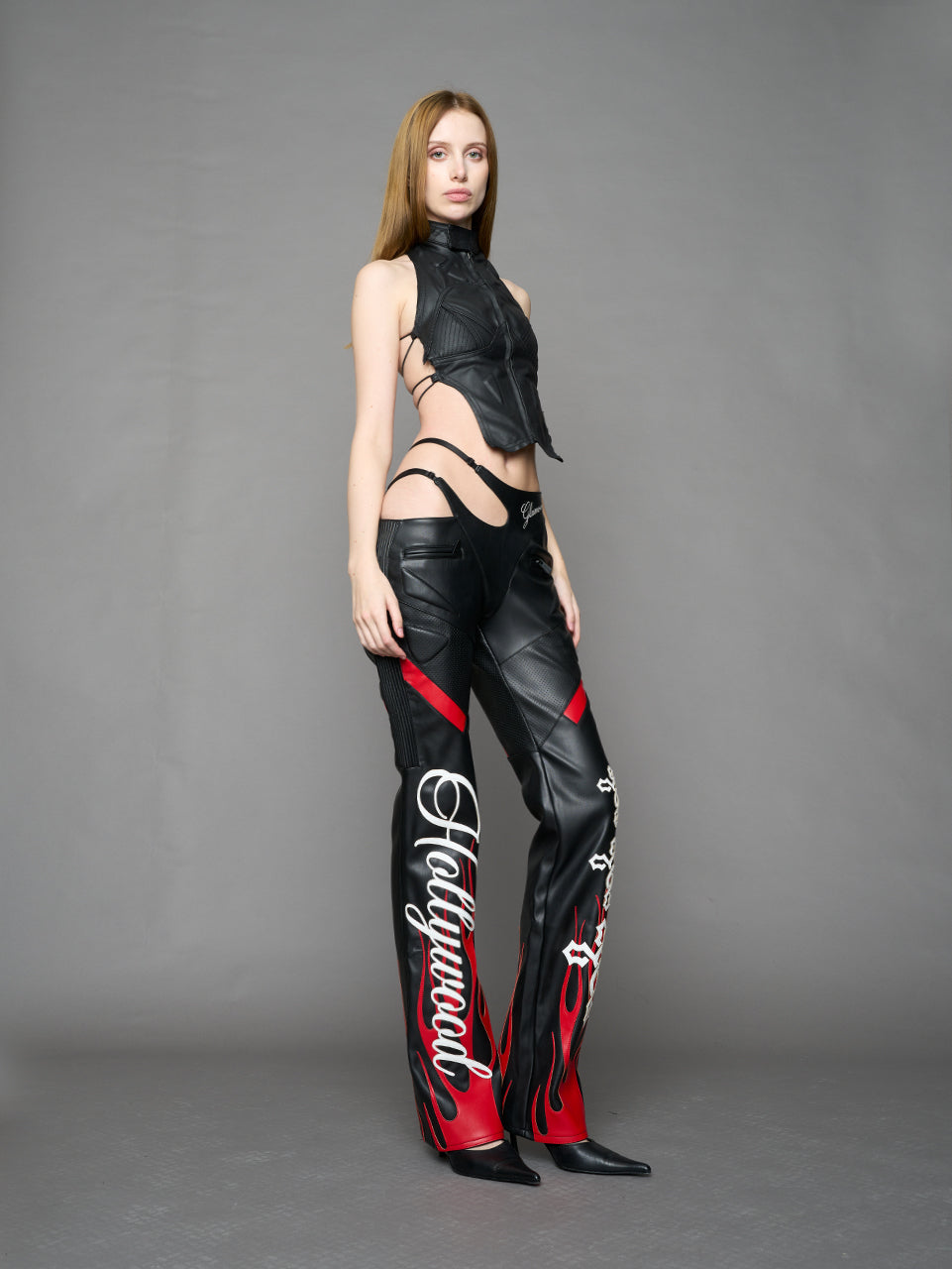 Model wears black/red vegan leather flare trousers with motocross paneling, padded details, flame and cross stitching, slogan embroidery, and an asymmetric waistband with adjustable elastic thong straps.