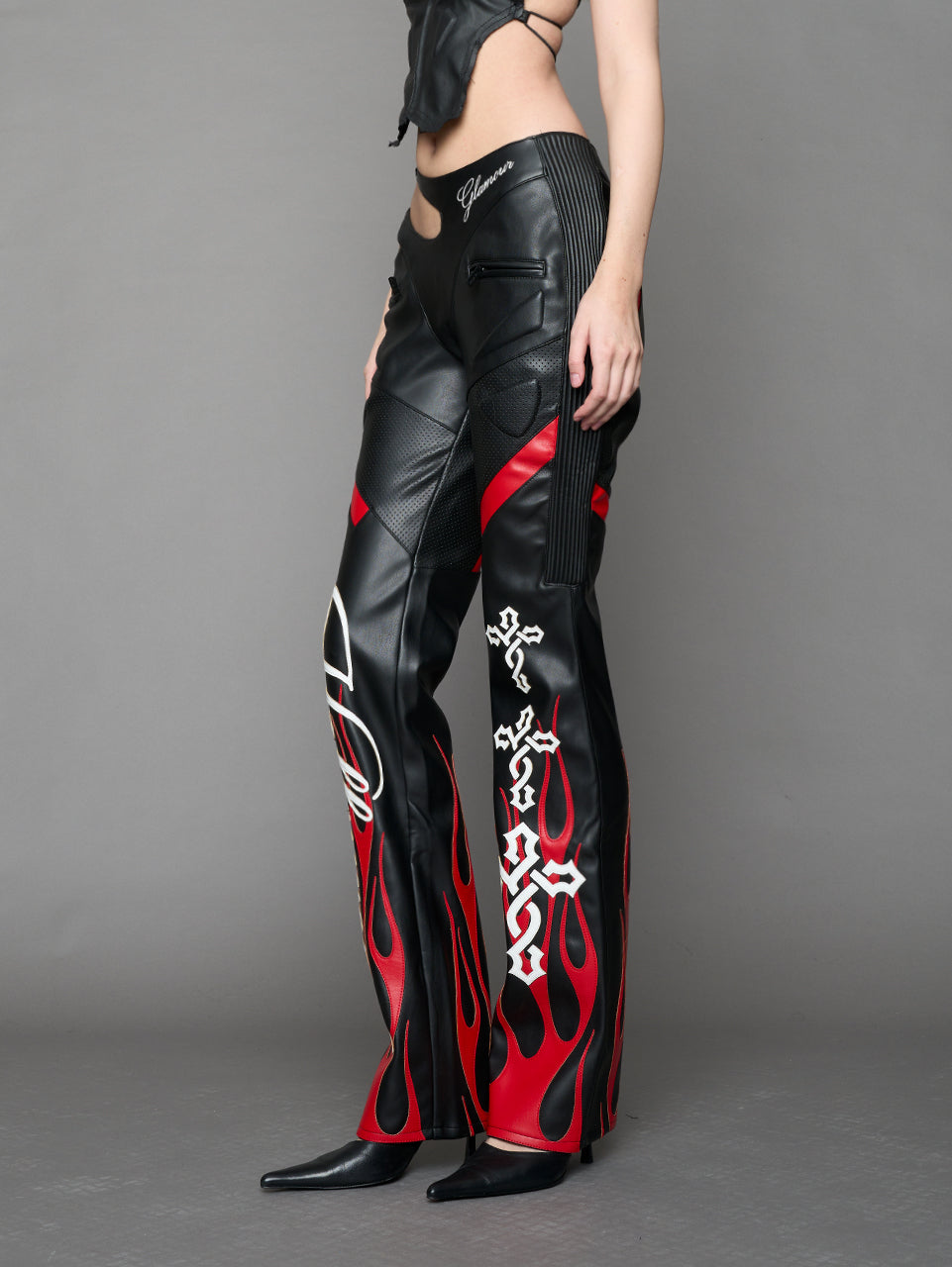Model wears black/red vegan leather flare trousers with motocross paneling, padded details, flame and cross stitching, slogan embroidery, and an asymmetric waistband with adjustable elastic thong straps.