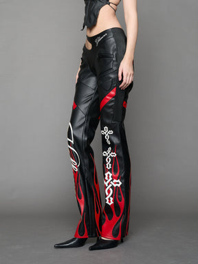 Model wears black/red vegan leather flare trousers with motocross paneling, padded details, flame and cross stitching, slogan embroidery, and an asymmetric waistband with adjustable elastic thong straps.