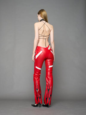 Model wears red vegan leather flare trousers with motocross paneling, padded details, flame and cross stitching, slogan embroidery, and an asymmetric waistband with adjustable elastic thong straps.