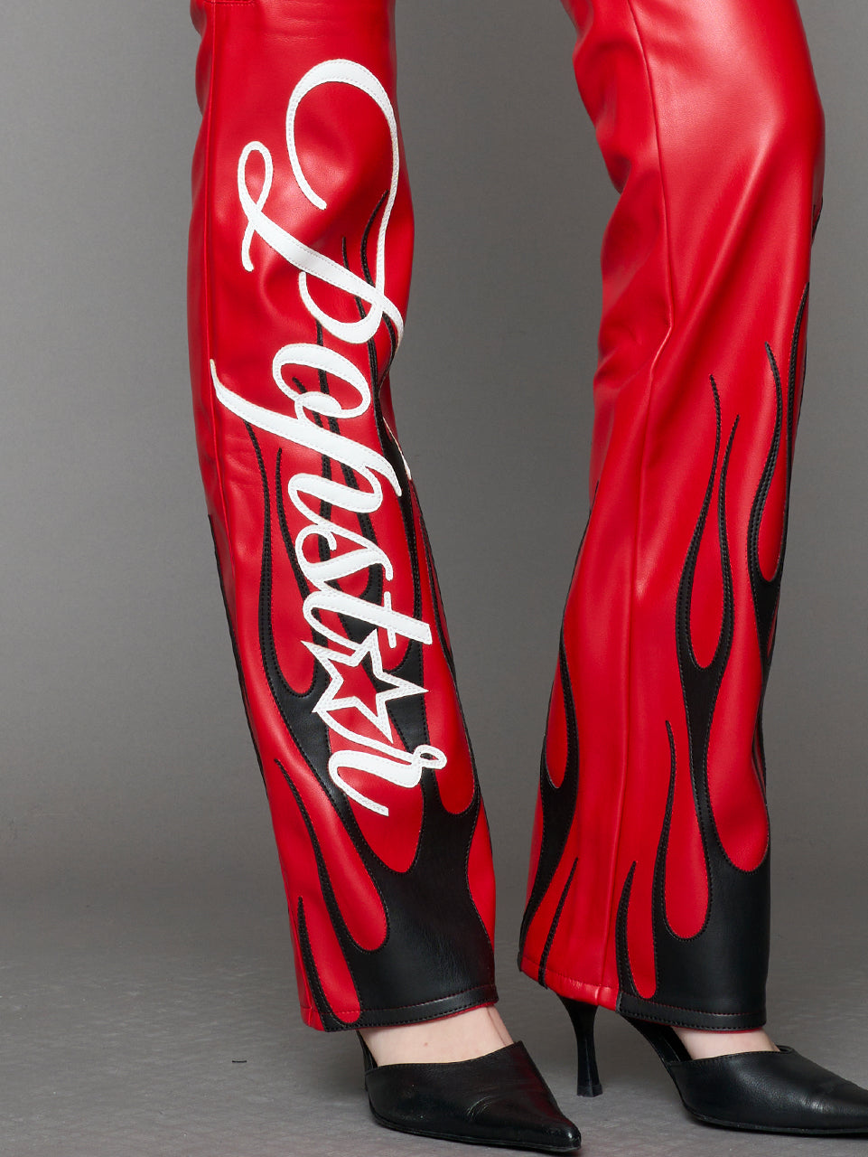 Model wears red vegan leather flare trousers with motocross paneling, padded details, flame and cross stitching, slogan embroidery, and an asymmetric waistband with adjustable elastic thong straps.