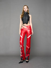Model wears red vegan leather flare trousers with motocross paneling, padded details, flame and cross stitching, slogan embroidery, and an asymmetric waistband with adjustable elastic thong straps.