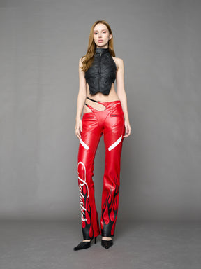 Model wears red vegan leather flare trousers with motocross paneling, padded details, flame and cross stitching, slogan embroidery, and an asymmetric waistband with adjustable elastic thong straps.