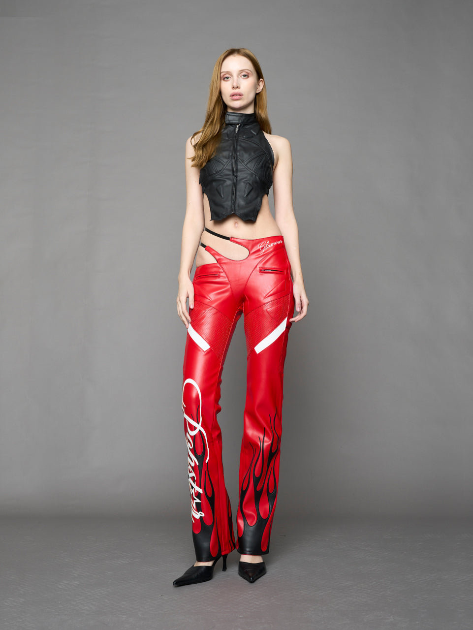 Model wears red vegan leather flare trousers with motocross paneling, padded details, flame and cross stitching, slogan embroidery, and an asymmetric waistband with adjustable elastic thong straps.