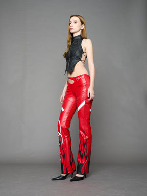 Model wears red vegan leather flare trousers with motocross paneling, padded details, flame and cross stitching, slogan embroidery, and an asymmetric waistband with adjustable elastic thong straps.