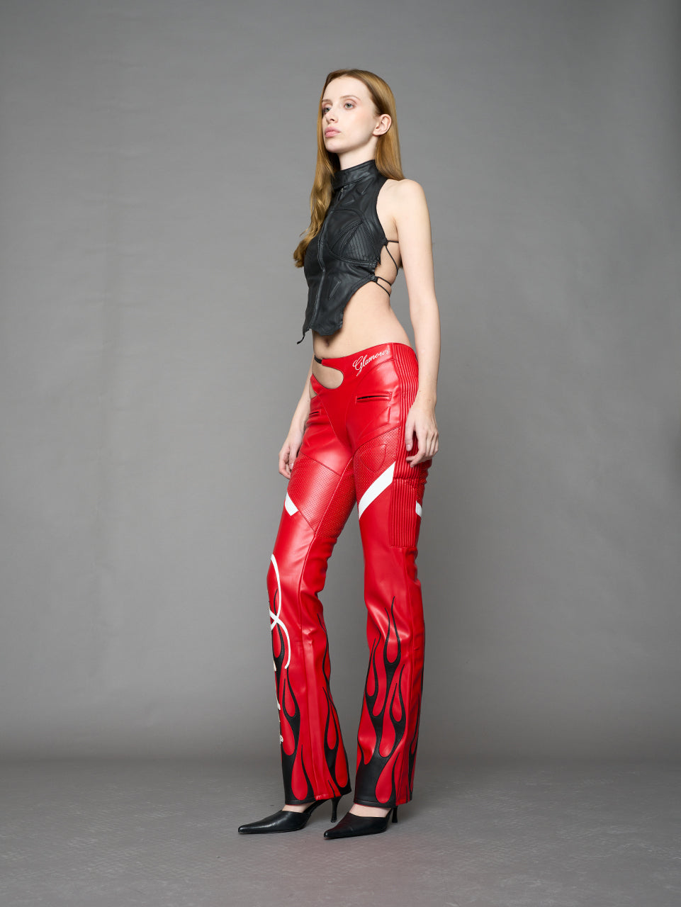 Model wears red vegan leather flare trousers with motocross paneling, padded details, flame and cross stitching, slogan embroidery, and an asymmetric waistband with adjustable elastic thong straps.