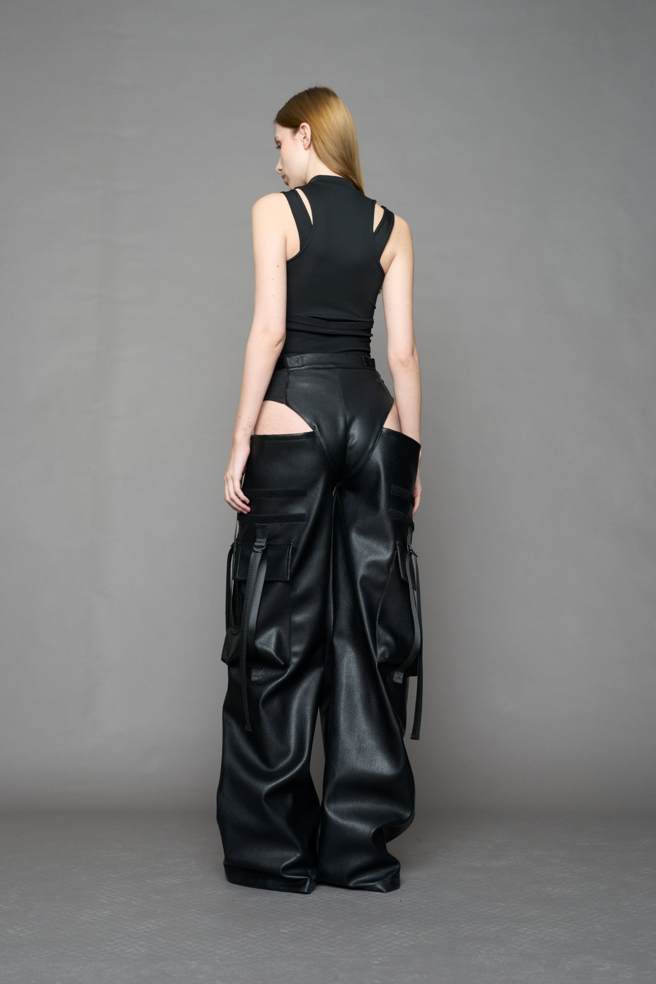 Model wearing Black vegan leather panty trousers featuring an integrated stretch panel for comfort, adjustable belt detail, and a relaxed high-waist silhouette.