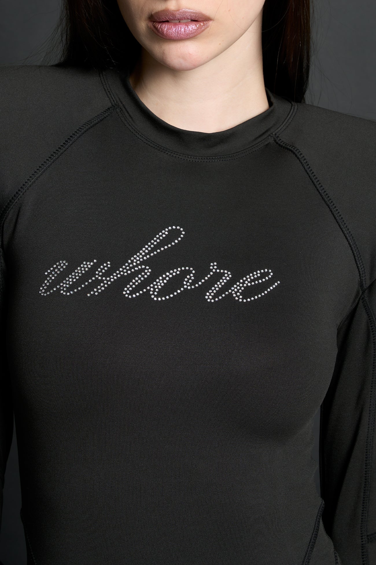 Model wears a long-sleeved black lycra stretch bodysuit with string back and detachable padded shoulders, rhinestoned slogan, thumbhole gloves.