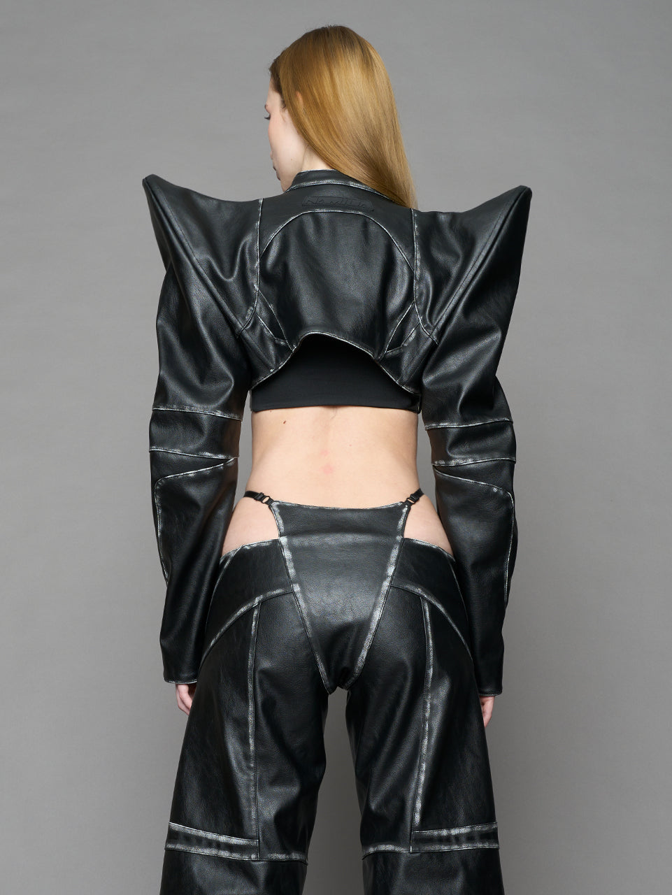 Model wearing a black vegan leather spike shoulder bolero jacket with cut-out design on a gray background