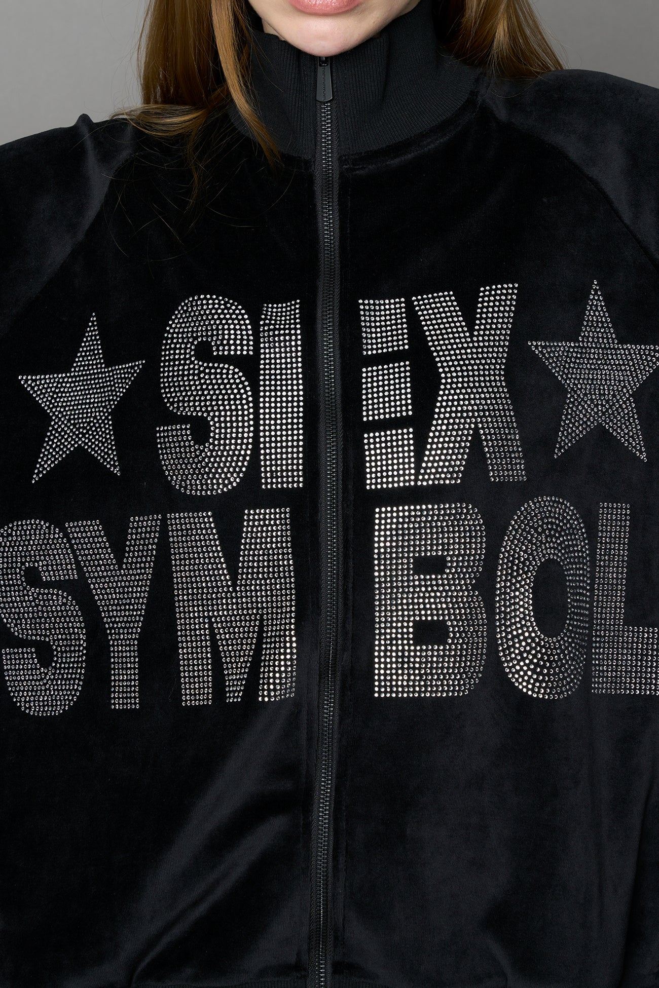 Person wearing a black velour jacket with 'S*X SYMBOL' crystal slogan on a gray background
