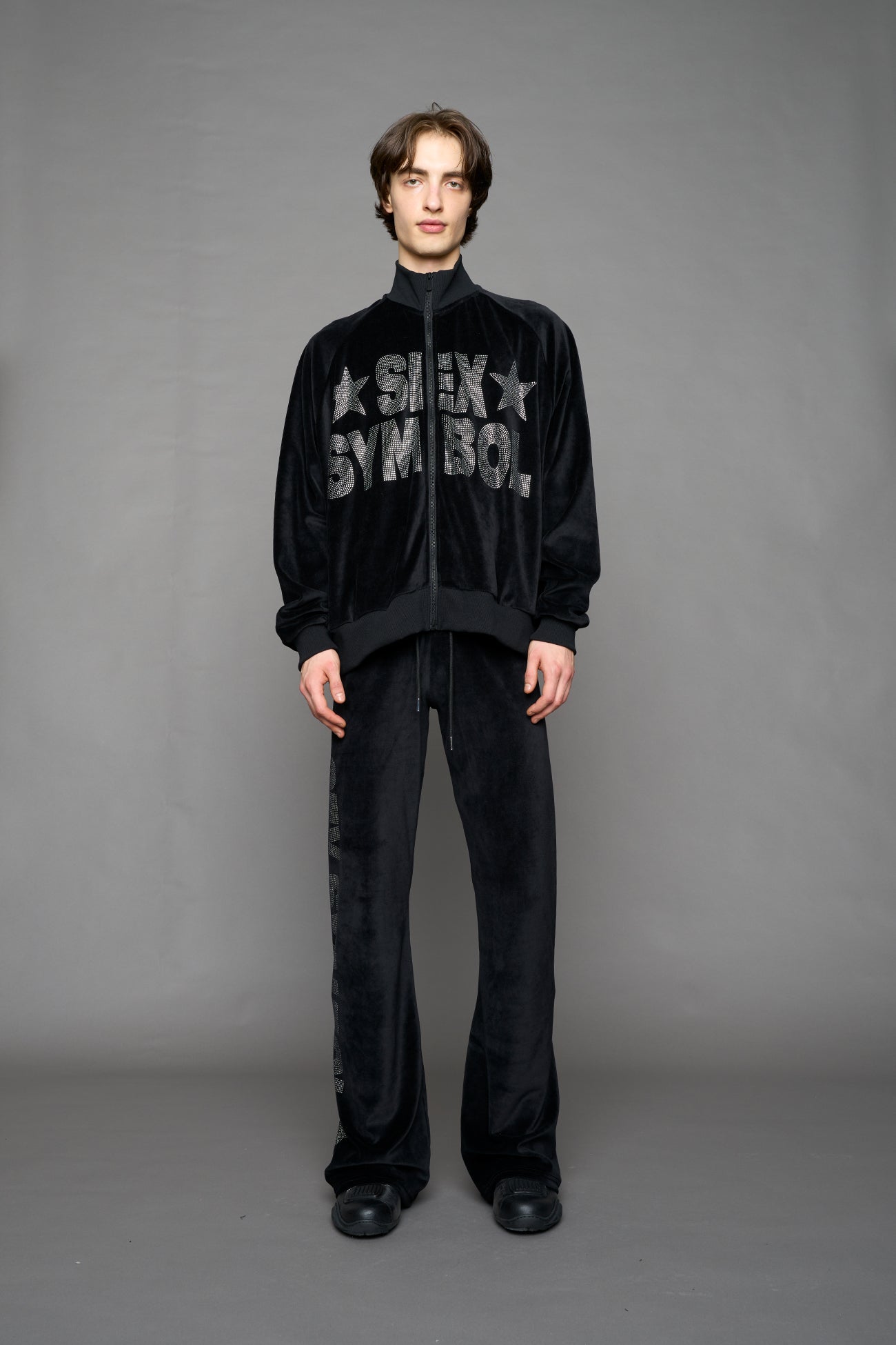 Person wearing a black velour jacket with 'S*X SYMBOL' crystal slogan on a gray background