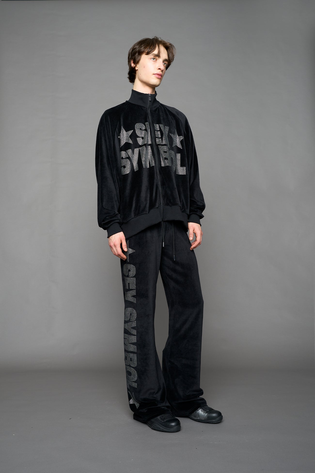 Person wearing a black velour jacket with 'S*X SYMBOL' crystal slogan on a gray background