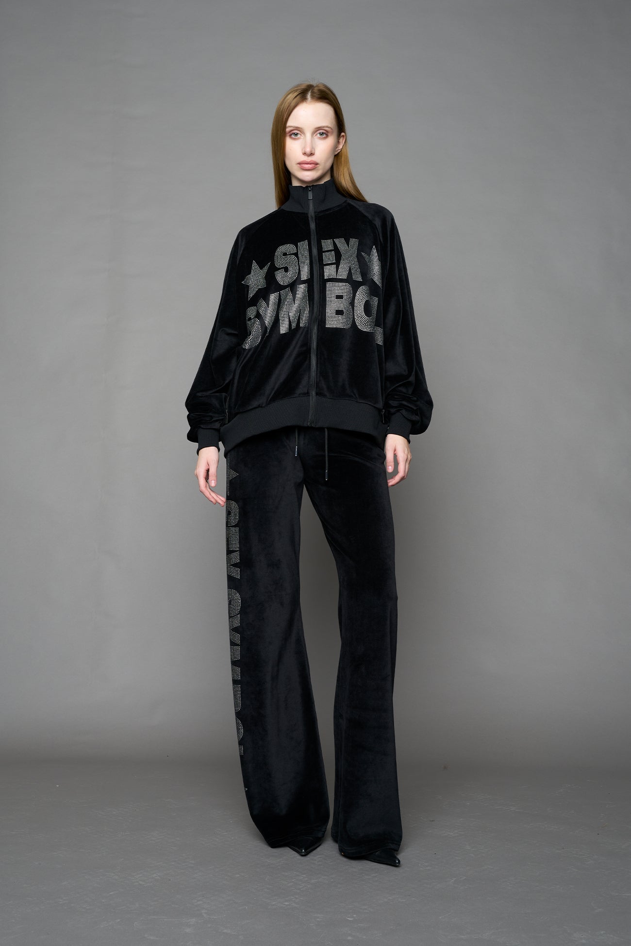 Person wearing a black velour jacket with 'S*X SYMBOL' crystal slogan on a gray background