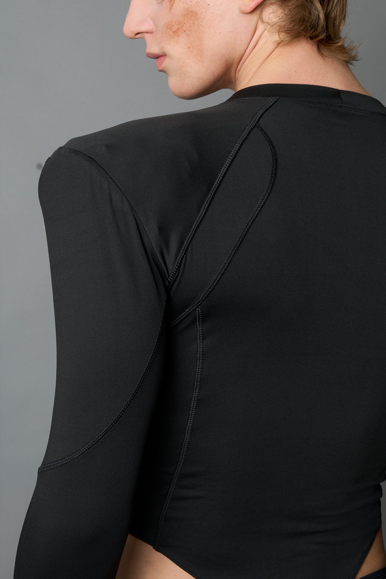 Male model wearing a black high-cut thong bodysuit with long sleeves and thumbhole details, edgy avant-garde fashion on a neutral grey studio background