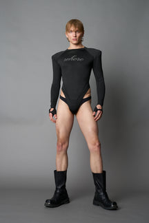 Male model wearing a black high-cut thong bodysuit with long sleeves and thumbhole details, edgy avant-garde fashion on a neutral grey studio background