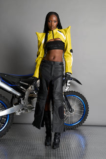 Model wears the yellow Namilia Spike Moto Bolero Jacket with wire shoulder pads, cropped hemline, branded front zipper, motocross-style lines, and black rubber logo tag on the back.
