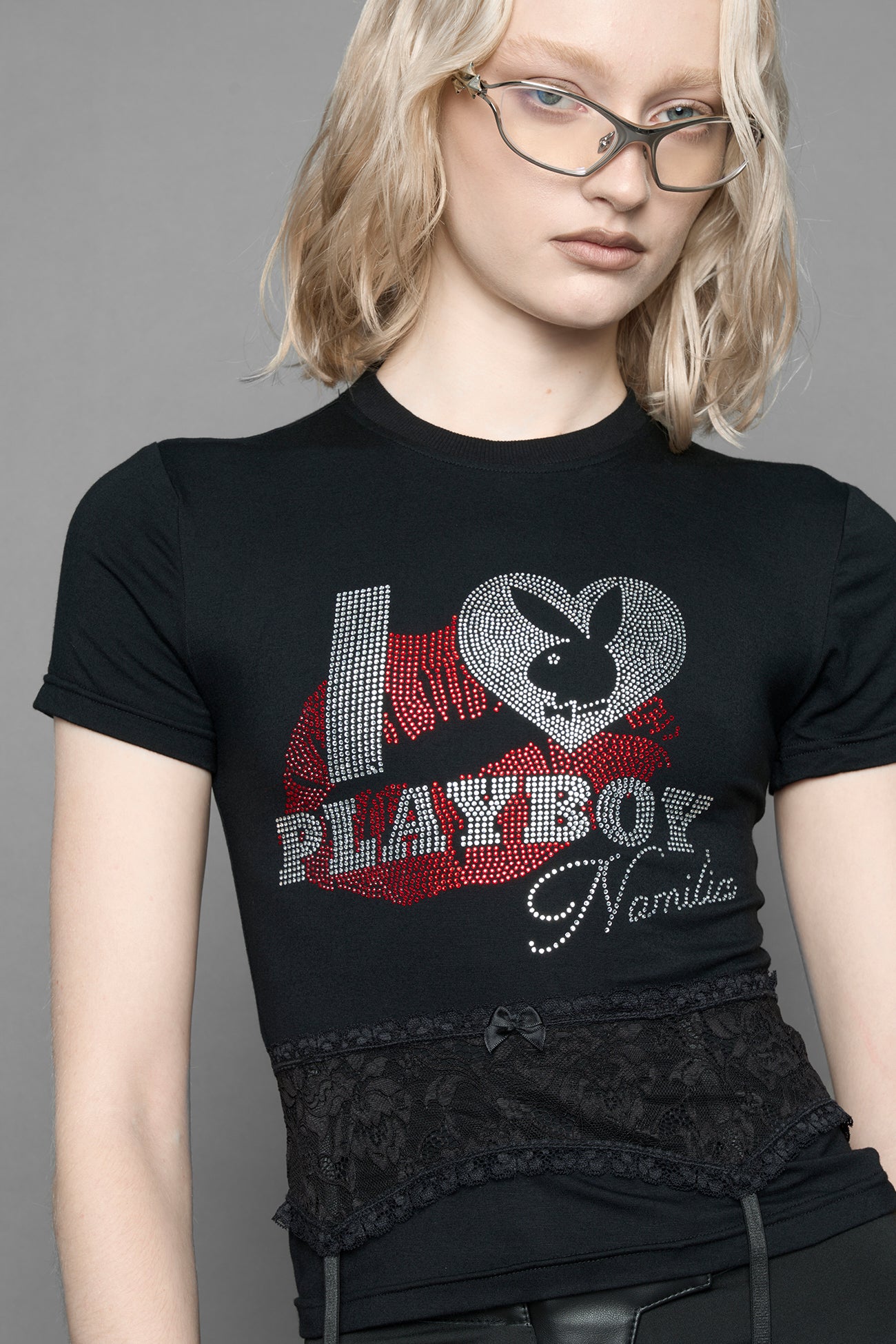 model wearing a black short-sleeve graphic t-shirt with rhinestone embroidery of red lip print and silver 'I <heart> PLAYBOY' design. T-Shirt has a lingerie black lace garter with suspender with metal silver Bunny Heads attached.