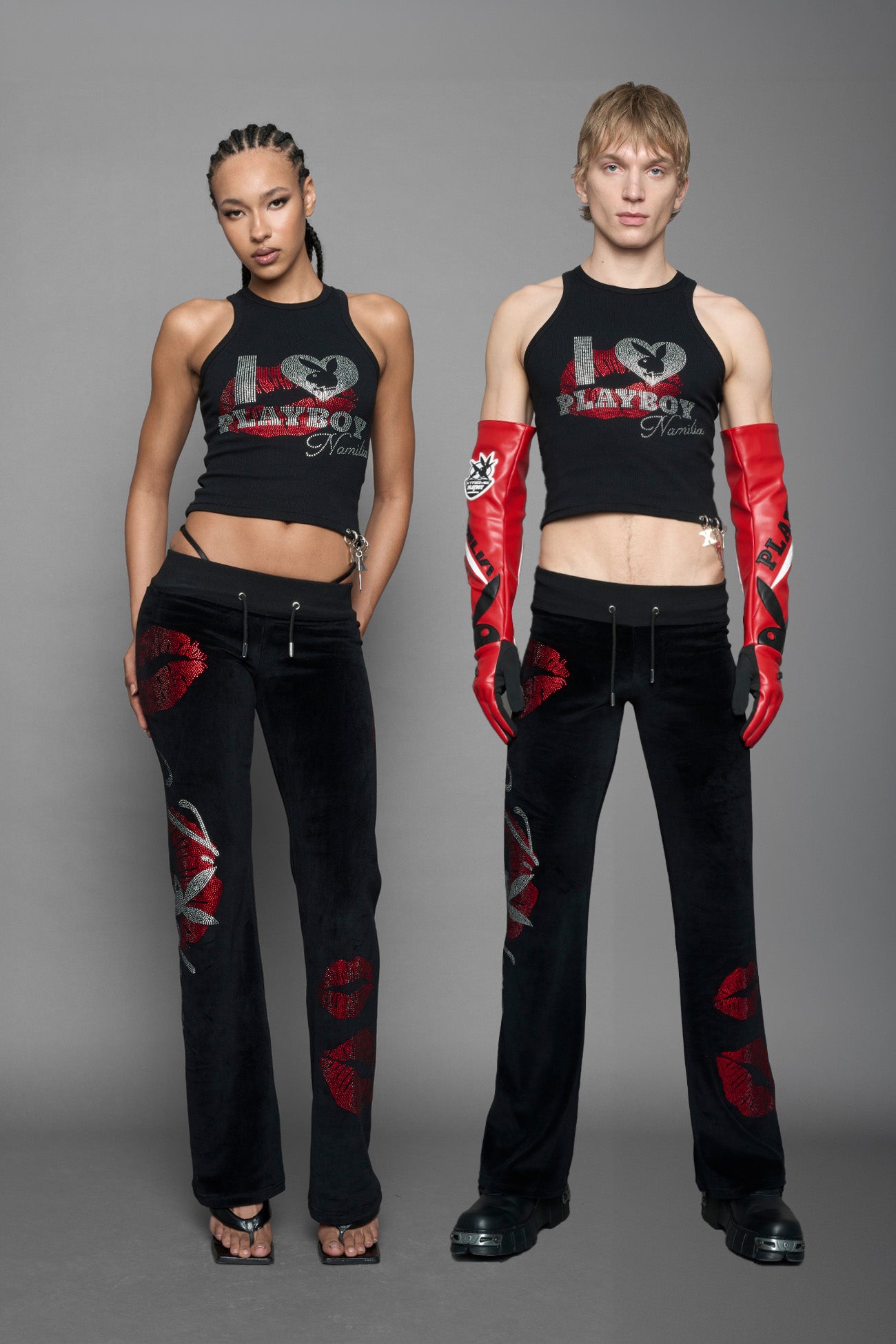 model wearing a black cotton tank top with a rhinestone embroidery of red and silver 'I <heart> PLAYBOY Namilia' graphic. At the hem on the wearers left side, there are three eyelets with detachable charms. Model is standing against a solid gray studio background.
