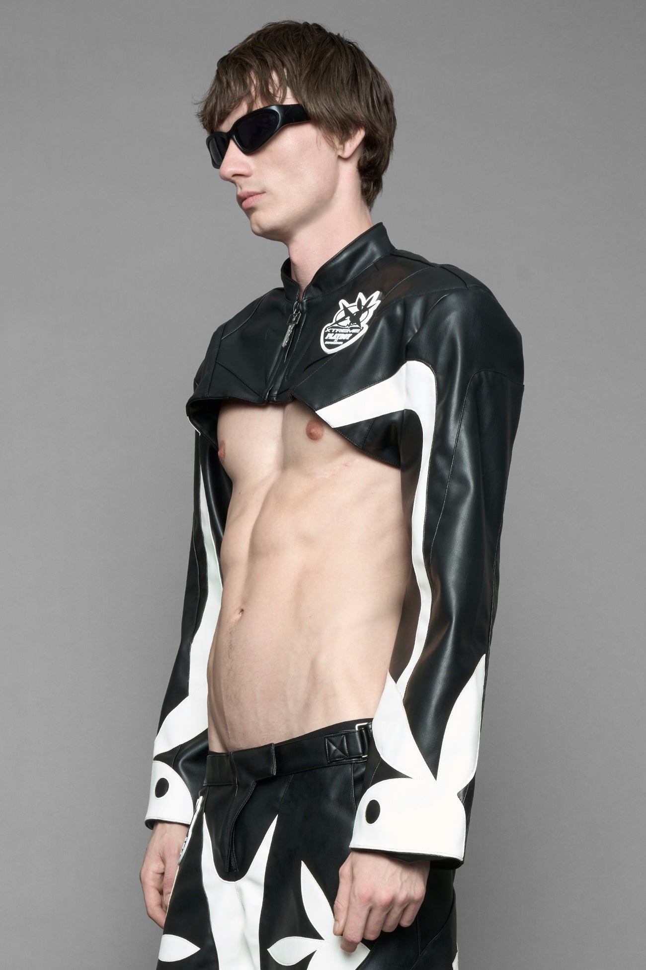model wearing a vegan leather cropped jacket with broad shoulders in black with white paneling. The jacket has PU leather embroidery of 'Namilia' and 'Playboy' with a key formed zipper pull and a rubbertag on the left breast.
