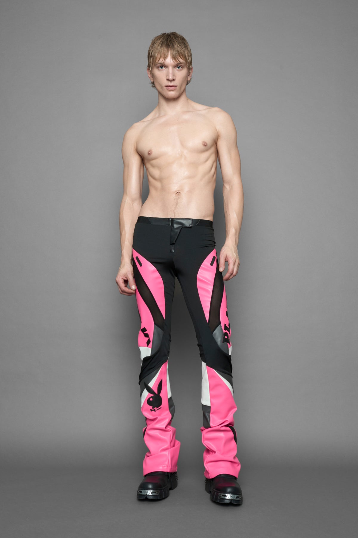 A female model wearing a pink and black form fitted Pants with PU Leather Embroidery of 'Namilia', 'Playboy'. The pants have black Mesh fabric and dark grey PU leather  panels and are slightly flared from knee to hem.