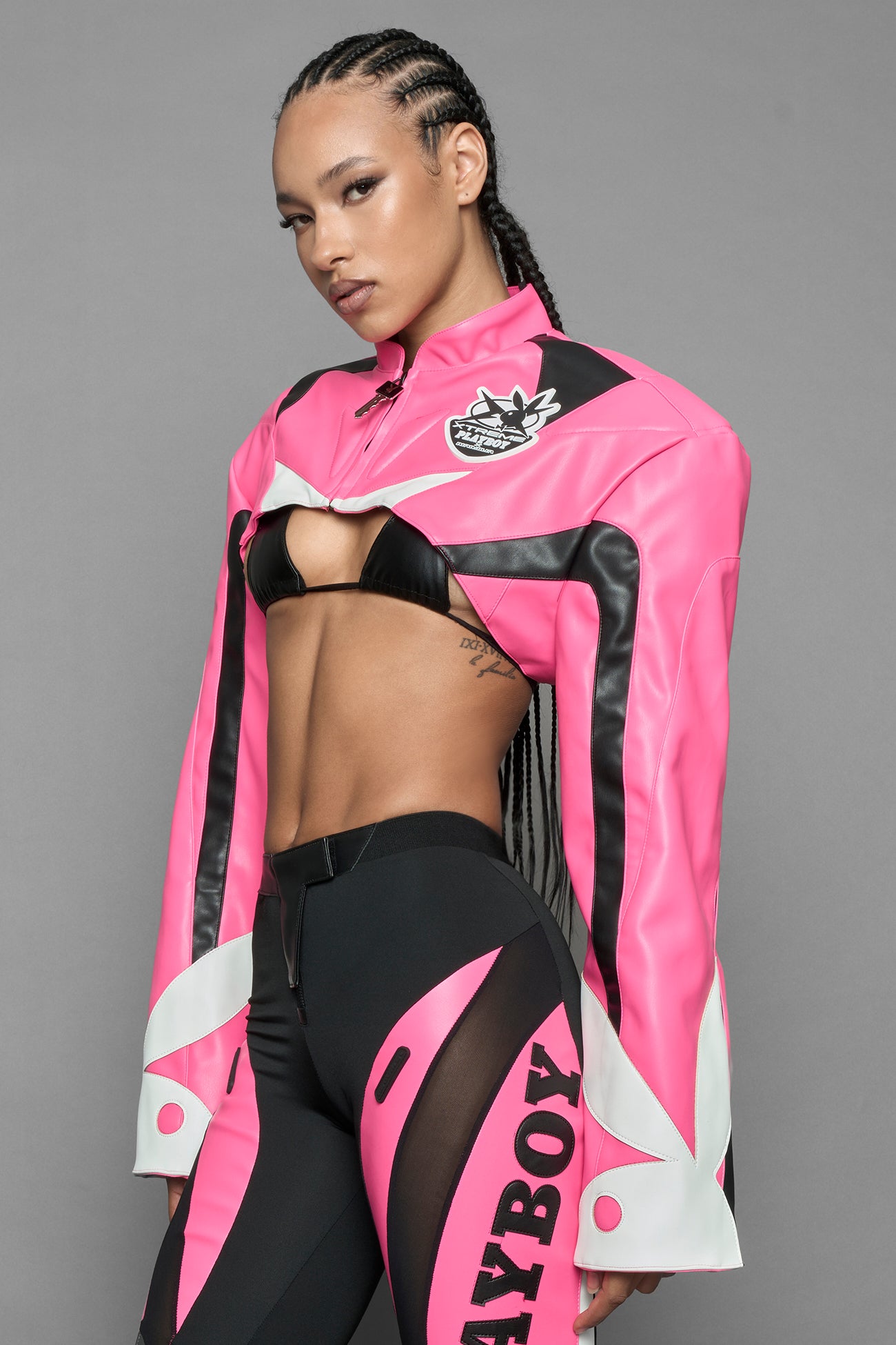 model wearing a vegan leather cropped jacket with broad shoulders in pink with black and white paneling. The jacket has PU leather embroidery of 'Namilia' and 'Playboy' with a key formed zipper pull and a rubbertag on the left breast.
