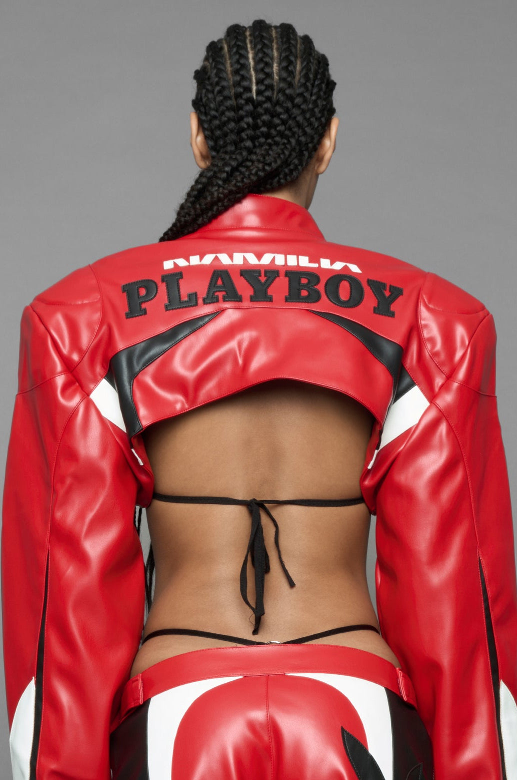 model wearing a vegan leather cropped jacket with broad shoulders in red with black and white paneling. The jacket has PU leather embroidery of 'Namilia' and 'Playboy' with a key formed zipper pull and a rubbertag on the left breast.
