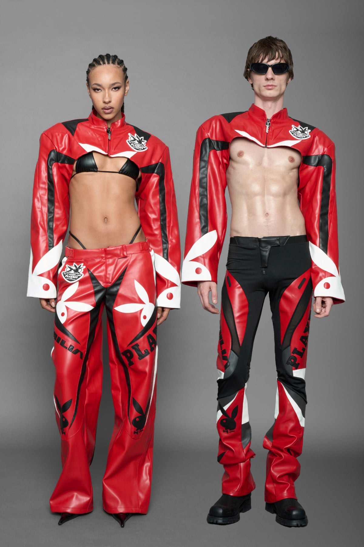 A male model wearing a vegan leather cropped jacket with broad shoulders in red with black and white paneling. The jacket has PU leather embroidery of 'Namilia' and 'Playboy' with a key formed zipper pull and a rubbertag on the left breast.