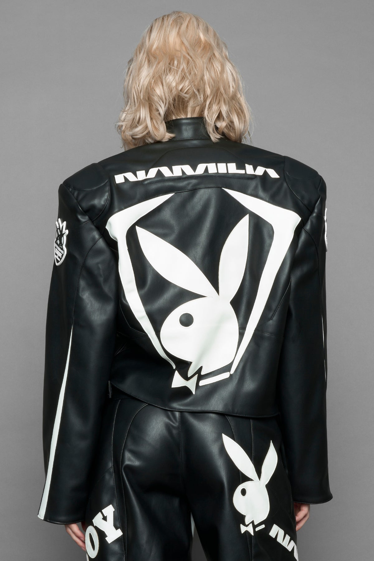 model wearing black vegan leather zip-up jacket with white PU leather 'Namilia' and 'Playboy' embroidery and black and white paneling. On the left chest there is a white Playboy Bunny Embroidery.