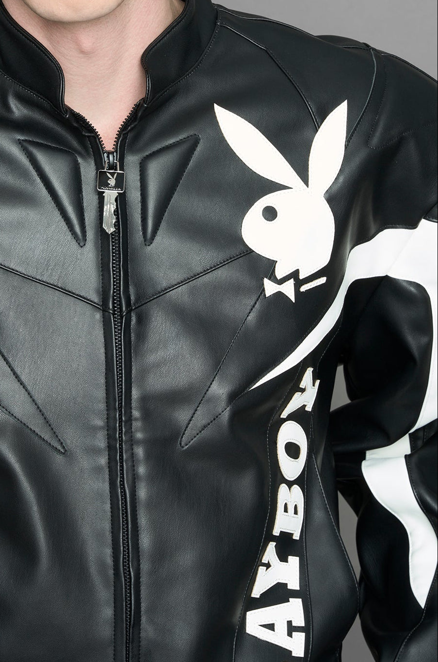 model wearing black vegan leather zip-up jacket with white PU leather 'Namilia' and 'Playboy' embroidery and black and white paneling. On the left chest there is a white Playboy Bunny Embroidery.