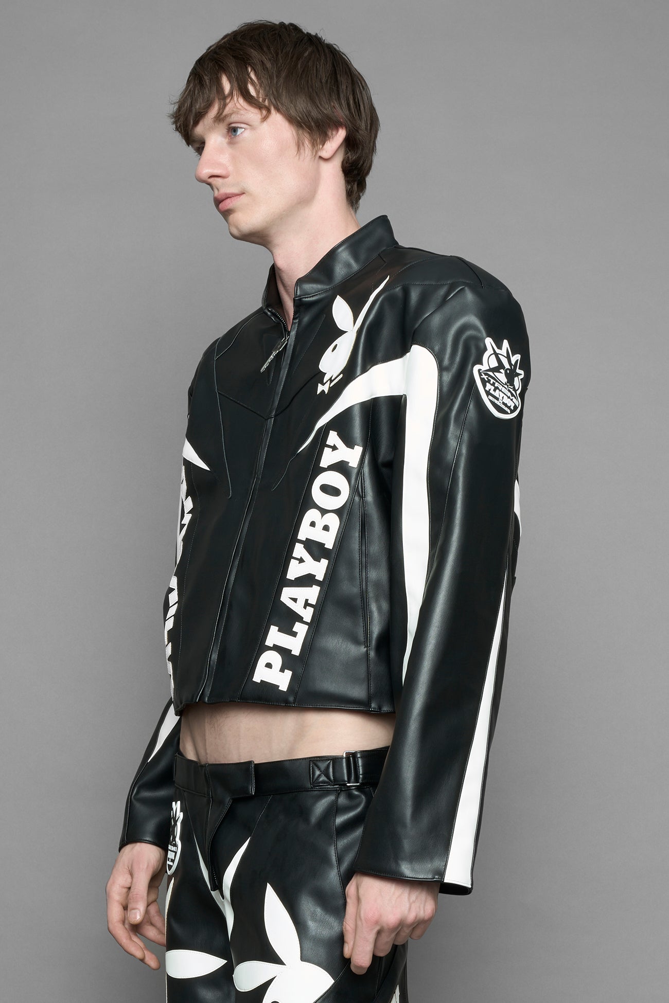 model wearing black vegan leather zip-up jacket with white PU leather 'Namilia' and 'Playboy' embroidery and black and white paneling. On the left chest there is a white Playboy Bunny Embroidery.