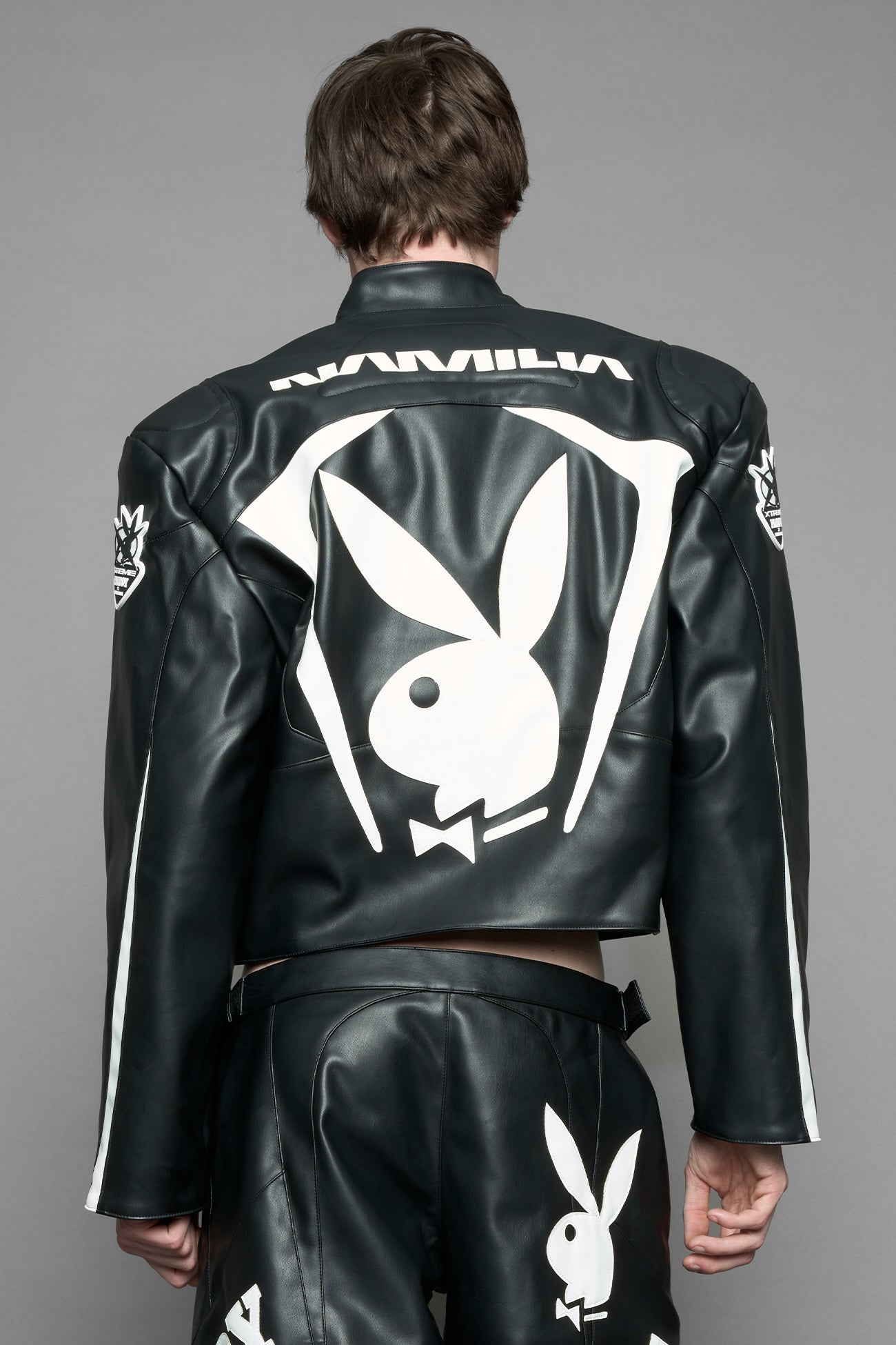 model wearing black vegan leather zip-up jacket with white PU leather 'Namilia' and 'Playboy' embroidery and black and white paneling. On the left chest there is a white Playboy Bunny Embroidery.