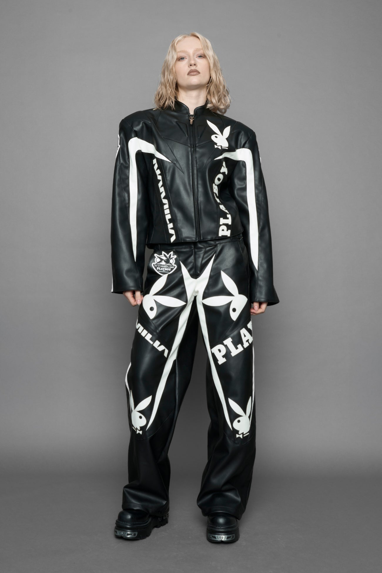 model wearing black vegan leather zip-up jacket with white PU leather 'Namilia' and 'Playboy' embroidery and black and white paneling. On the left chest there is a white Playboy Bunny Embroidery.