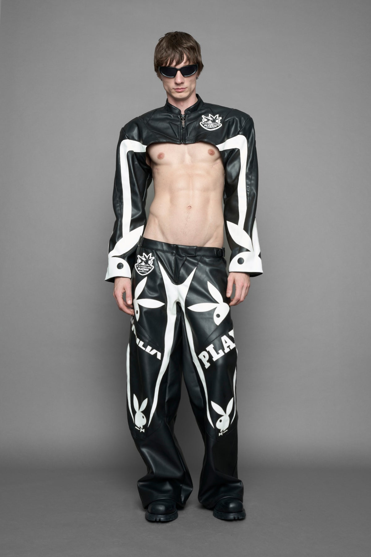 model wearing a vegan leather cropped jacket with broad shoulders in black with white paneling. The jacket has PU leather embroidery of 'Namilia' and 'Playboy' with a key formed zipper pull and a rubbertag on the left breast.