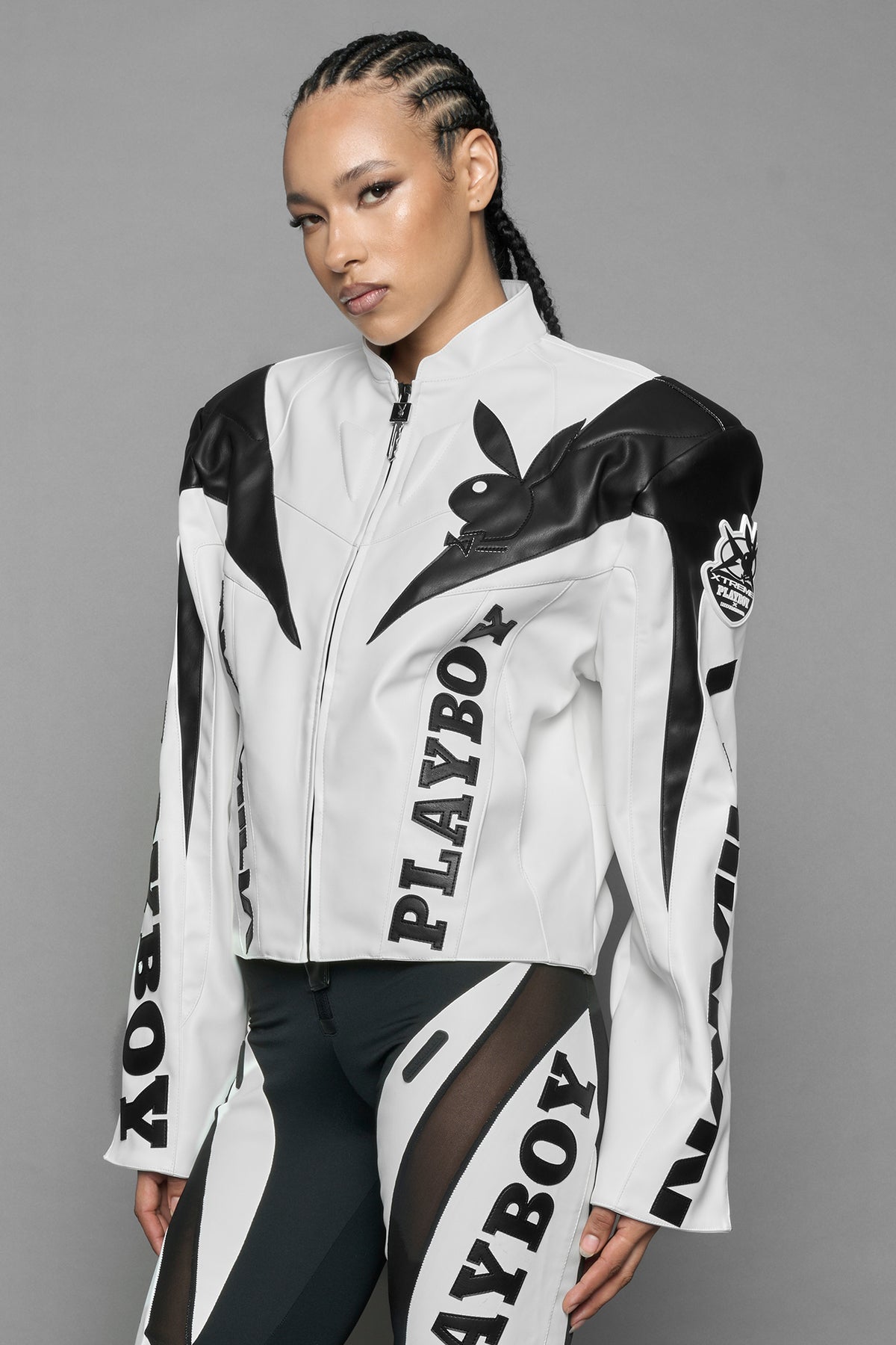 model wearing white vegan leather zip-up jacket with black PU leather 'Namilia' and 'Playboy' embroidery and black and white paneling. On the left chest there is a white Playboy Bunny Embroidery.