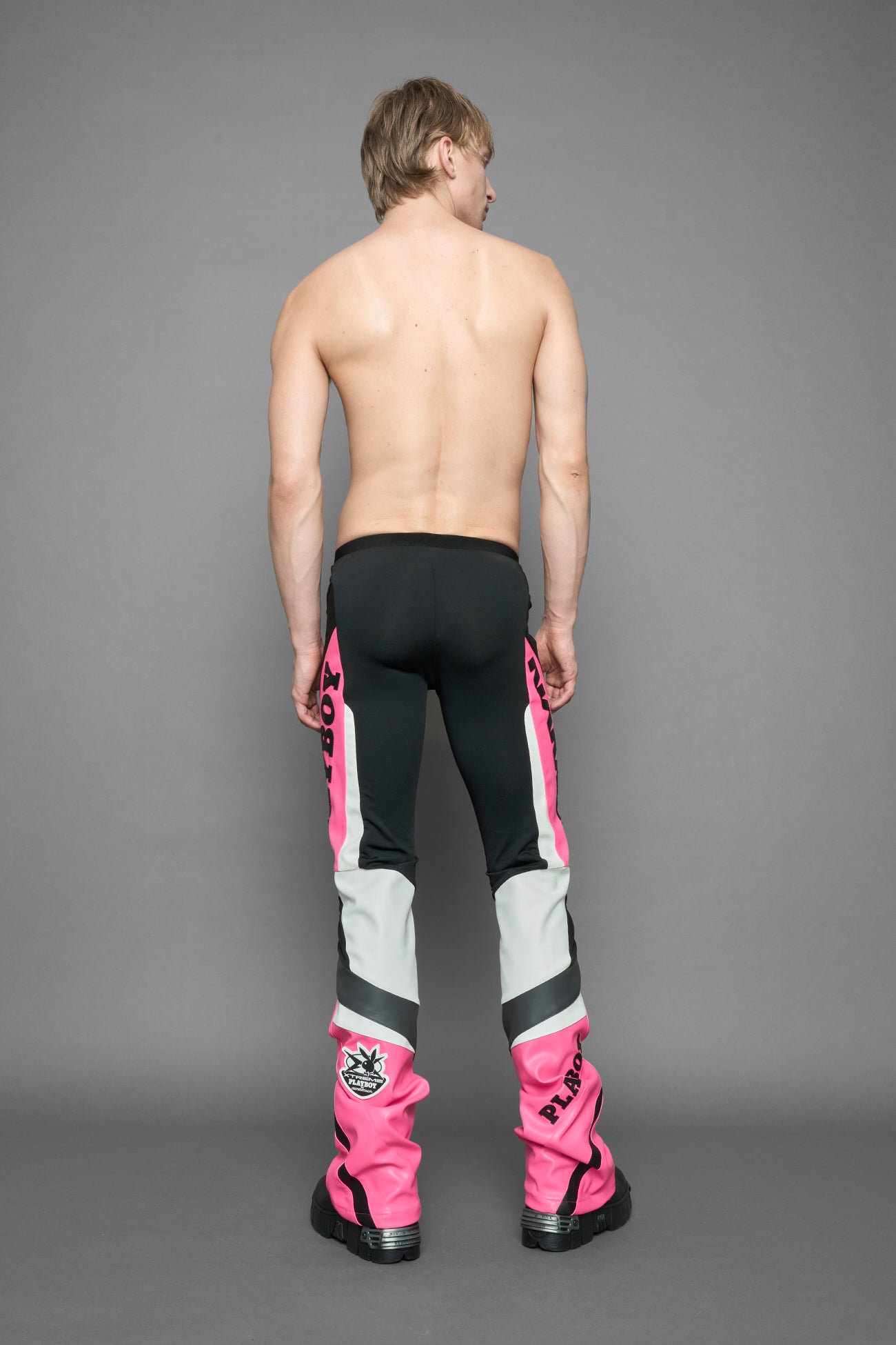 A female model wearing a pink and black form fitted Pants with PU Leather Embroidery of 'Namilia', 'Playboy'. The pants have black Mesh fabric and dark grey PU leather  panels and are slightly flared from knee to hem.