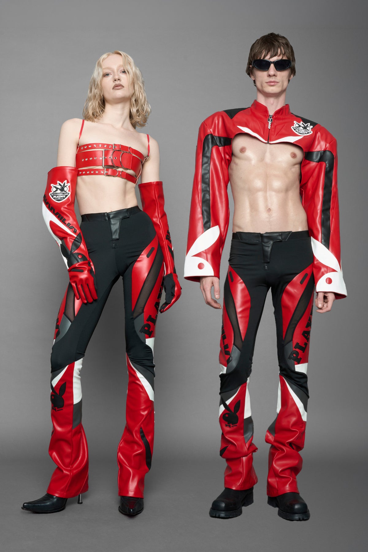 model wearing a red and black form fitted Pants with PU Leather Embroidery of 'Namilia', 'Playboy'. The pants have black Mesh fabric and dark grey PU leather  panels and are slightly flared from knee to hem