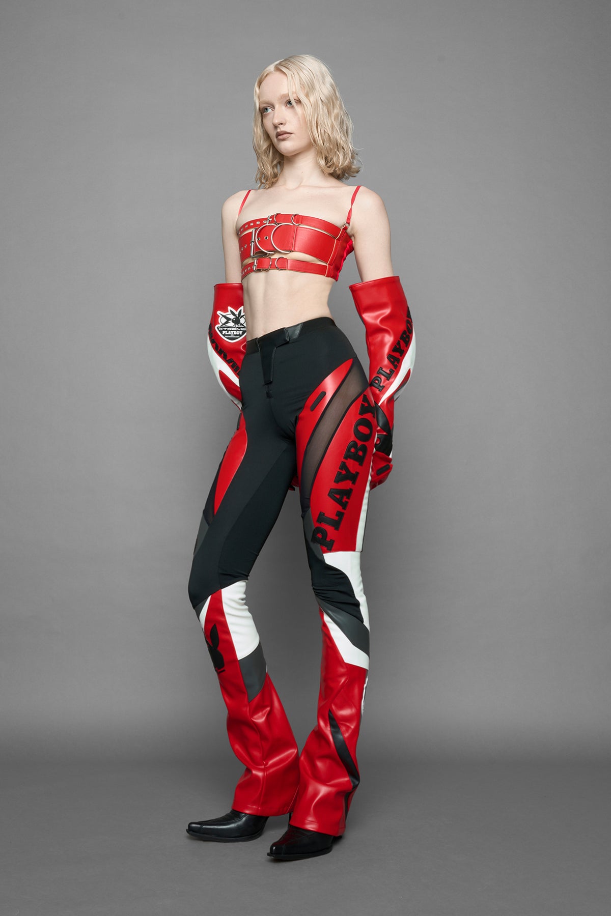 model wearing a red and black form fitted Pants with PU Leather Embroidery of 'Namilia', 'Playboy'. The pants have black Mesh fabric and dark grey PU leather  panels and are slightly flared from knee to hem