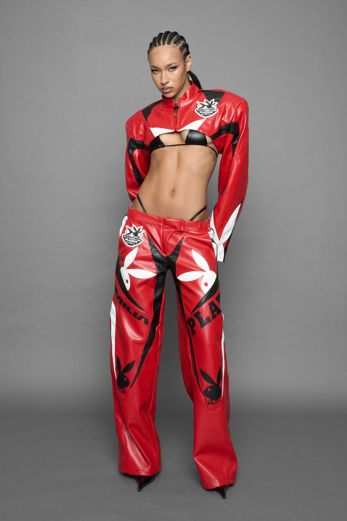 model wearing a vegan leather cropped jacket with broad shoulders in red with black and white paneling. The jacket has PU leather embroidery of 'Namilia' and 'Playboy' with a key formed zipper pull and a rubbertag on the left breast.