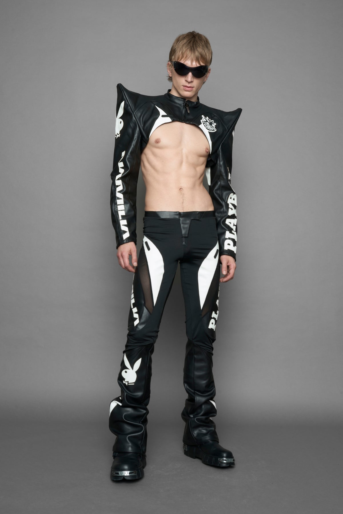  model wearing a vegan leather cropped jacket with spike silhouette shoulders in black with white paneling. The jacket has PU leather embroidery of 'Namilia' and 'Playboy' with a key formed zipper pull and a rubbertag on the left breast.