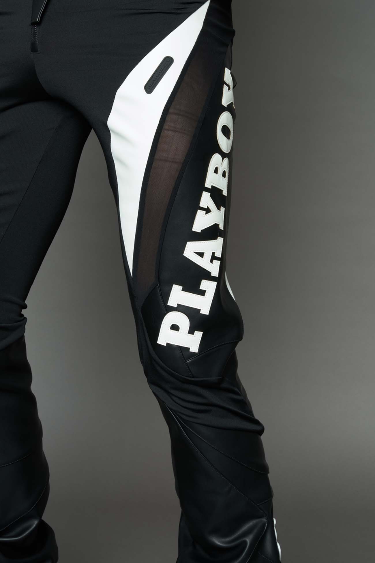 model wearing a black and white form fitted Pants with PU Leather Embroidery of 'Namilia', 'Playboy'. The pants have black Mesh fabric panels and are slightly flared from knee to hem