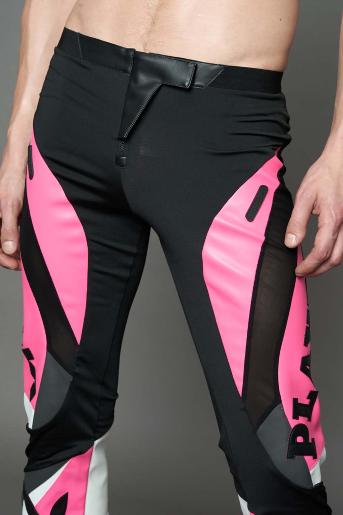 A female model wearing a pink and black form fitted Pants with PU Leather Embroidery of 'Namilia', 'Playboy'. The pants have black Mesh fabric and dark grey PU leather  panels and are slightly flared from knee to hem.