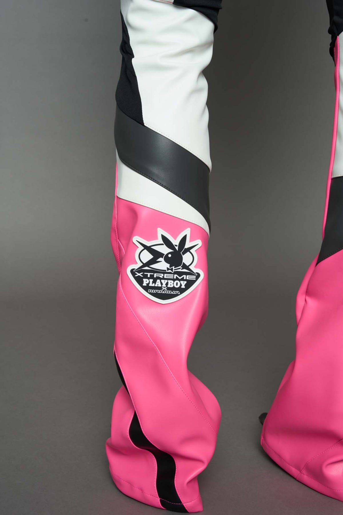 A female model wearing a pink and black form fitted Pants with PU Leather Embroidery of 'Namilia', 'Playboy'. The pants have black Mesh fabric and dark grey PU leather  panels and are slightly flared from knee to hem.